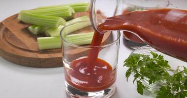 derramando orgânico recentemente espremido tomate suco para dentro uma vidro. salsão e salsinha em uma branco mesa. saudável e vegetariano Comida conceito. ultra 4k video