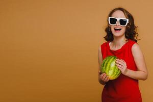 A beautiful girl in glasses and a red dress holds a watermelon in her hands photo