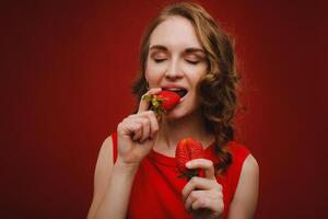 un hermosa niña en un rojo vestir en un rojo antecedentes sostiene un fresa en su manos y sonrisas foto