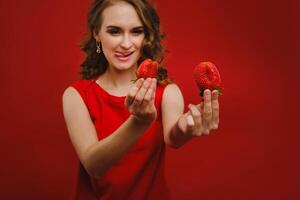 A beautiful girl in a red dress on a red background holds a strawberry in her hands and smiles photo