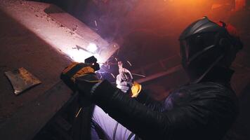 joven mecánico con un soldadura máquina en un antiguo sucio garaje a noche. hombre en proteccion haciendo soldadura trabajar, pasatiempo video