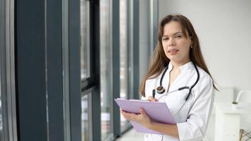ung caucasian kvinna läkare bär vit medicinsk täcka och stetoskop med dokument i henne händer ser på kamera. leende kvinna läkare Framställ i sjukhus kontor video
