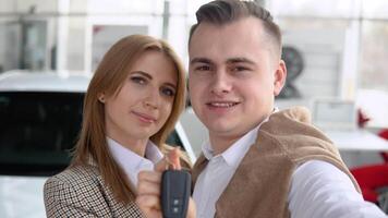 gelukkig paar nemen een selfie Bij een auto handel met een nieuw auto in de achtergrond. Mens en vrouw tonen sleutels van zijn nieuw auto in handel video