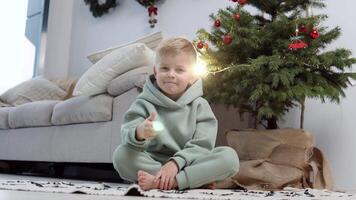 un rubio chico en un elegante casual traje se sienta en el alfombra cerca el Navidad árbol y puntos su dedo súper video