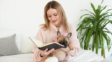 une Jeune femme dans une confortable poudre rose chandail et blanc bas est assis sur le lit avec sa chien et lit une livre video