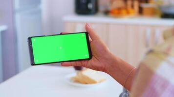 Woman watching a video on phone with green screen in kitchen during morning. Green screen chroma mock up isolated mockup background ready to be replaced with your text, logo or advertisement. Using internet, social networks and browsing