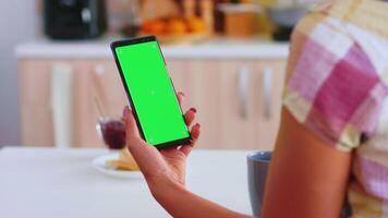 Woman looking at phone with green touchscreen while drinking a cup of coffee in kitchen. Green screen chroma mock up isolated mockup background ready to be replaced with your text, logo or advertisement. Using internet, social networks and browsing video