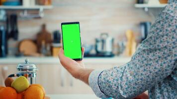 Mature man holding smartphone with green screen during breakfast in kitchen. Elderly person with chroma key isolated mock-up mockup for easy replacement video