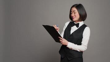 Asian hotel concierge creating a checklist of registrations, working on online reservations and bookings in studio. Receptionist looking at clipboard papers over grey background. Camera A. video