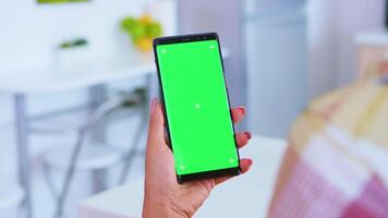 Woman watching video content on phone with green screen in kitchen. Green screen chroma mock up isolated mockup background ready to be replaced with your text, logo or advertisement. Using internet, social networks and browsing