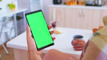 mujer participación portátil teléfono con verde pantalla mientras Bebiendo café en cocina. verde pantalla croma burlarse de arriba aislado Bosquejo antecedentes Listo a ser sustituido con tu texto, logo o anuncio publicitario. utilizando Internet, social redes y hojeada video