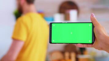 Woman hand holding smartphone with green screen in kitchen while husband is in the background. Green screen chroma mock up isolated mockup background ready to be replaced with your text, logo or advertisement. Using internet, social networks and browsing video