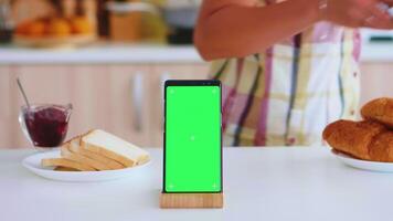 Close up of phone with green screen in kitchen while woman taking a cup of coffee. Green screen chroma mock up isolated mockup background ready to be replaced with your text, logo or advertisement. Using internet, social networks and browsing video