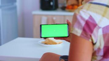 femme en train de regarder une téléphone avec vert écran dans cuisine tandis que en buvant une tasse de café. vert écran chrominance moquer en haut isolé maquette Contexte prêt à être remplacé avec votre texte, logo ou publicité. en utilisant l'Internet, social réseaux et navigation video