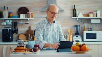Senior parent browsing on tablet pc in kitchen during breakfast having a healthy lifestyle. Elderly person with tablet portable pad PC in retirement age using mobile apps, modern internet online information technology with touchscreen video