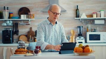 mayor padre hojeada en tableta ordenador personal en cocina durante desayuno teniendo un sano estilo de vida. mayor persona con tableta portátil almohadilla ordenador personal en Jubilación años utilizando móvil aplicaciones, moderno Internet en línea información tecnología con pantalla táctil video