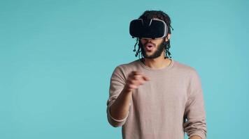 middle eastern man wearing virtual reality headset, doing swiping gestures. BIPOC person using high tech futuristic modern VR gadget, isolated over blue studio background, camera A video