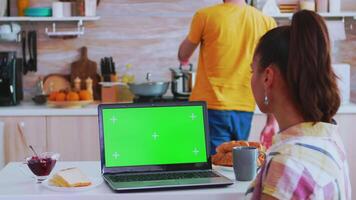 donna Guardando il computer portatile con verde croma chiave e marito preparazione prima colazione. verde schermo croma finto su isolato modello sfondo pronto per essere sostituito con il tuo testo, logo o annuncio. utilizzando Internet, sociale reti e navigazione video