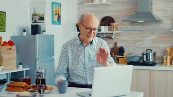 Senior Mann chatten und winken während ein Video Anruf mit Laptop im Küche halten ein Tasse von Kaffee. Alten Person mit Internet online Plaudern Technologie Video Webcam Herstellung ein Video Anruf Verbindung Kamera Kommunikation Konferenz Anruf
