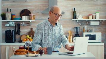 Sénior homme travail sur portable dans cuisine pendant petit déjeuner profiter une tasse de café. personnes âgées retraité la personne travail de maison, télétravail en utilisant éloigné l'Internet emploi en ligne la communication sur moderne La technologie carnet video