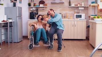 alegre discapacitado mujer en silla de ruedas tomando un selfie con marido en cocina. esperanzado marido con minusválido discapacitado invalidez inválido parálisis Desventaja persona siguiente a a él, Ayudar su a reintegrar video