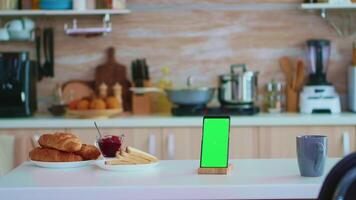 Mobile phone with chroma key in kitchen while housewife is cooking in the background. Green screen chroma mock up isolated mockup background ready to be replaced with your text, logo or advertisement. Using internet, social networks and browsing video