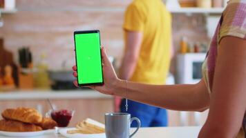 Wife using phone with green screen in kitchen while husband is making breakfast. Green screen chroma mock up isolated mockup background ready to be replaced with your text, logo or advertisement. Using internet, social networks and browsing video