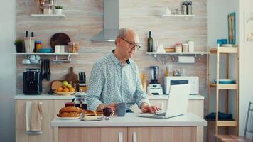 Daily life of senior man in kitchen during breakfast using laptop holding a cup of coffee. Elderly retired person working from home, telecommuting using remote internet job online communication on modern technology notebook video