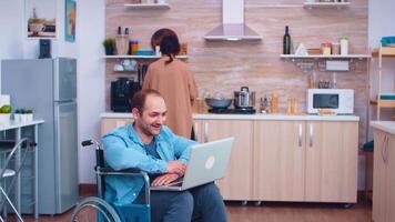 Disabled man waving on video conference using laptop in kitchen. Corporate man with paralysis handicap disability handicapped difficulties working after accident having internet online video call conference communication for professional use.