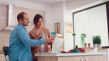 casal sorridente às computador portátil tela dentro cozinha. esposa segurando copo do café. marido e esposa cozinhando receita Comida. feliz saudável juntos estilo de vida. família procurando para conectados refeição. saúde fresco salada video