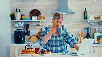 mayor mujer de negocios Bebiendo café y trabajando en ordenador portátil en cocina durante desayuno. mayor retirado persona trabajando desde hogar, teletrabajo utilizando remoto Internet trabajo en línea comunicación en moderno tecnología cuaderno video