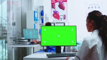 dokter gebruik makend van computer met groen scherm mockup in ziekenhuis en assistent wandelingen uit van kabinet vervelend blauw uniform. medisch in wit jas werken Aan toezicht houden op met chroma sleutel in kliniek kabinet naar controleren geduldig diagnostiseren. video