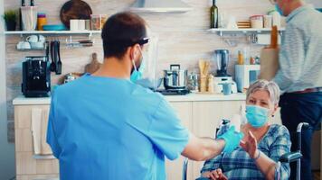 Handicapped senior woman in wheelchair wearing face mask discussing with doctor about treatment during coronavirus pandemic and home visit. Social worker offering pills to handicapped elderly woman. Geriatrician helping prevent covid-19 spread video