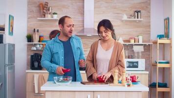 man het zingen Aan rood papier terwijl vrouw is snijdend een ander een voor salade gebruik makend van houten bijl in keuken. Koken voorbereidingen treffen gezond biologisch voedsel gelukkig samen levensstijl. vrolijk maaltijd in familie met groenten video