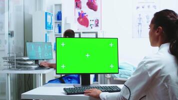 Assistant giving doctor patient x-ray while she is working on computer with copy space green screen available in hospital cabinet. Medic in white coat working on monitor with chroma key in clinic cabinet to check patient diagnose. video