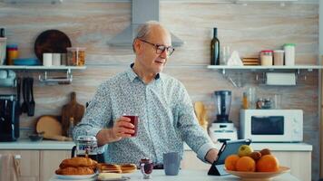 Senior Mann mit Tablette Computer während Trinken Tee während Frühstück im Küche. Alten Person mit Tablette tragbar Pad pc im Pensionierung Alter mit Handy, Mobiltelefon Apps, modern Internet online Information Technologie mit Berührungssensitiver Bildschirm video