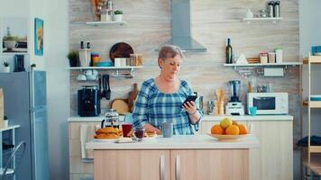 mayor mujer hojeada en móvil en cocina durante desayuno y sonriente a el cámara. auténtico mayor persona utilizando moderno teléfono inteligente Internet tecnología. en línea comunicación conectado a el mundo video