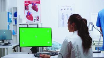 Doctor taking notes on computer with chroma key green screen and nurse in blue uniform in hospital cabinet. Medic in white coat working on monitor with blank space in clinic cabinet to check patient diagnose. video