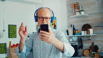 Happy mature adult listening music wearing headphones in kitchen during breakfast. Elderly retired person enjoying modern fun happy lifestyle, dancing relaxed, smiling and using modern technology video