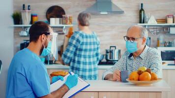 Social assistance with face mask and visor taking notes on clipboard while taking with senior man during home visit. Male nurse at retired couple visit explaining covid-19 spreading, help for risk group video