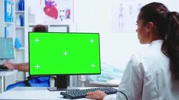 médico escrevendo diagnosticar em computador com verde tela e assistente vestindo azul uniforme dentro a fundo. médico dentro branco casaco trabalhando em monitor com croma chave dentro clínica gabinete para Verifica paciente diagnosticar. video