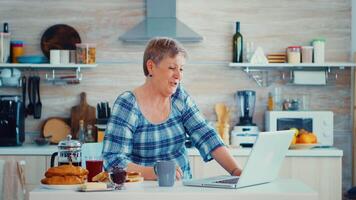 Senior Frau Trinken Kaffee und winken während ein Video Konferenz mit Familie im Küche während haben Frühstück. Alten Person mit Internet online Plaudern Technologie Video Webcam Herstellung ein Video Anruf Verbindung Kamera Kommunikation Konferenz Anruf