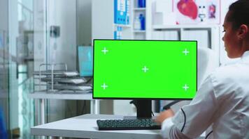 Doctor sits at computer with blank green screen display in hospital cabinet and assistant in blue uniform holding x-ray. Medic in white coat working on monitor with chroma key in clinic cabinet to check patient diagnose. video