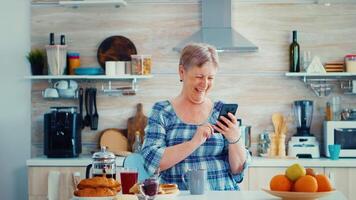 Senior mulher acenando durante vídeo conferência usando Smartphone dentro cozinha enquanto tendo café da manhã. idosos pessoa usando Internet conectados bate-papo tecnologia vídeo Webcam fazer uma vídeo ligar conexão Câmera comunicação conferência ligar video