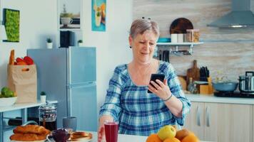 anziano donna utilizzando mobile aggeggio nel il cucina. autentico anziano persona utilizzando moderno smartphone Internet tecnologia. in linea comunicazione collegato per il mondo, anziano tempo libero tempo con aggeggio a la pensione età video