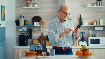 pensionista escuchando música en móvil teléfono mientras disfrutando un taza de café durante desayuno en cocina. contento relajado mayor mayor baile, divertido estilo de vida con moderno tecnología video