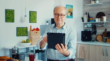 Happy older man in kitchen during breakfat using tablet pc. Elderly person with tablet portable pad PC in retirement age using mobile apps, modern internet online information technology with touchscreen video