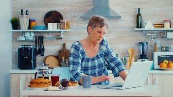 anziano donna ricerca in linea ricetta utilizzando il computer portatile nel cucina durante prima colazione. anziano pensionato persona Lavorando a partire dal casa, telelavoro utilizzando a distanza Internet lavoro in linea comunicazione su moderno tecnologia taccuino video