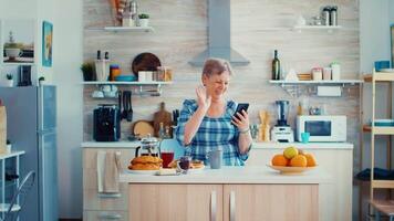Senior Frau Sprichwort Hallo während haben ein Video Anruf mit Familie mit Smartphone im Küche während Frühstück. Alten Person mit Internet online Plaudern Technologie Video Webcam Herstellung ein Video Anruf Verbindung Kamera Kommunikation Konferenz Anruf