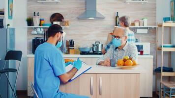 Doctor with face mask and visor writing prescription on clipboard during patient home visit. Male nurse social worker at retired senior couple visit explaining covid-19 spreading, help for people in risk group video